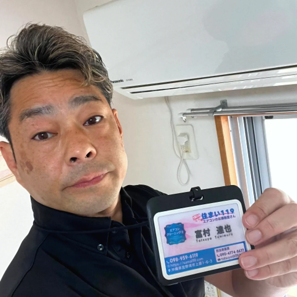 エアコンの前で住まい119の名札を掲げる富村達也